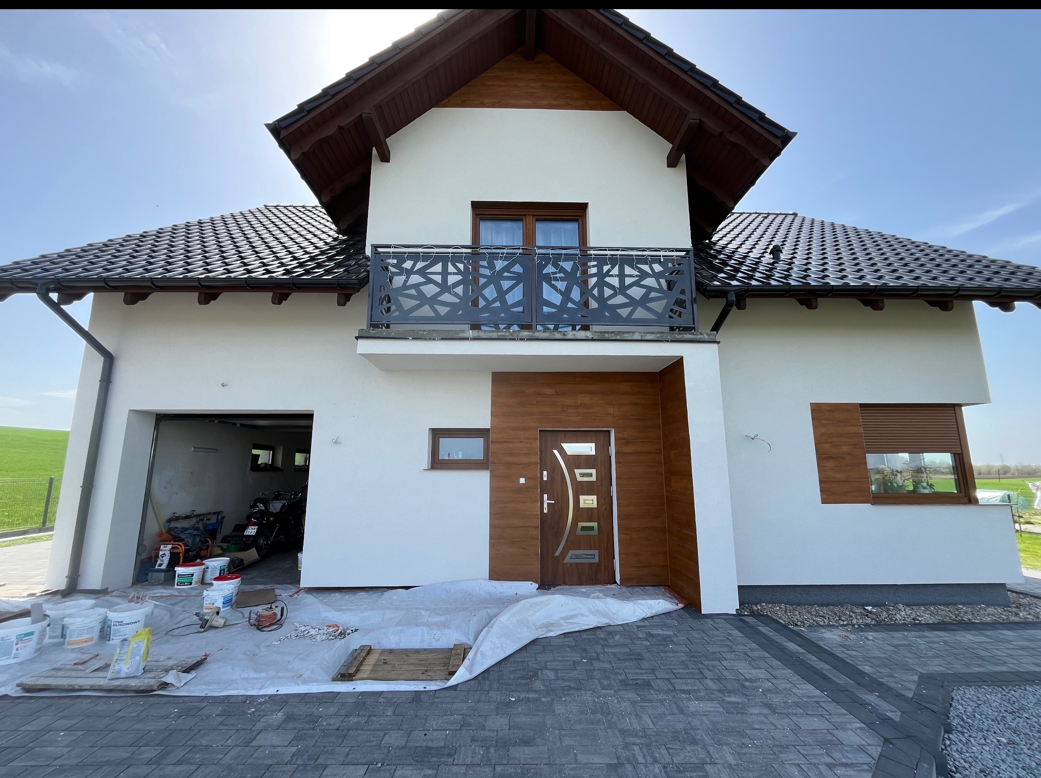 Elewacja domu jednorodzinnego po malowaniu, z widoczną nową stolarką drzwiową i okienną. Na pierwszym planie zabezpieczona folią kostka brukowa i farby.
