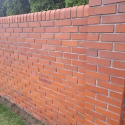 Nowy, czerwony mur z cegły klinkierowej z dekoracyjnym wykończeniem, widoczny na tle zielonych krzewów. Estetyczne ogrodzenie posesji.