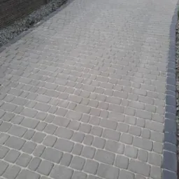 Ułożona szara kostka brukowa na podjeździe, obrzeża z ciemniejszych kostek, widoczne kamienie dekoracyjne i fragment ogrodzenia.