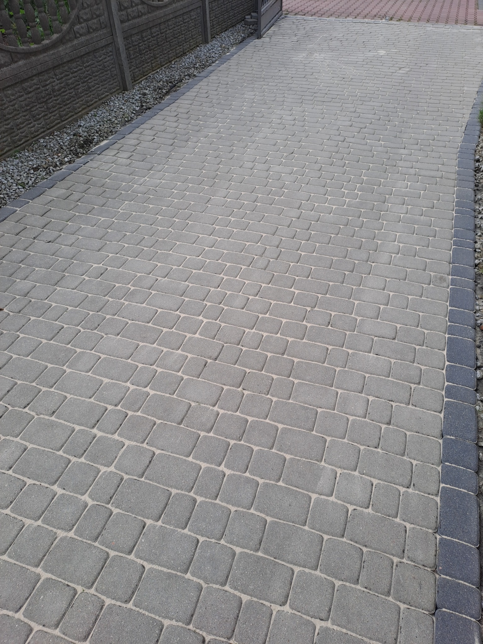 Ułożona szara kostka brukowa na podjeździe, obrzeża z ciemniejszych kostek, widoczne kamienie dekoracyjne i fragment ogrodzenia.