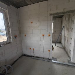 QWL - Wnętrze w trakcie prac elektrycznych: widoczne kable w peszlach prowadzone po ścianach z betonu komórkowego, puszki instalacyjne. Surowy stan, okno z widokiem na zewnątrz.