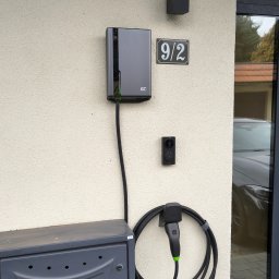 QWL - Szara stacja ładowania pojazdów elektrycznych GC zamontowana na elewacji domu obok skrzynki gazowej i numeru 9/2, z podłączonym kablem i wtyczką. Widoczny fragment samochodu.