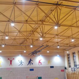 QWL - Oświetlenie hali sportowej z widoczną konstrukcją dachu, lampami i ścianą z kolorowymi sylwetkami sportowców. Na podłodze bramka do piłki ręcznej.