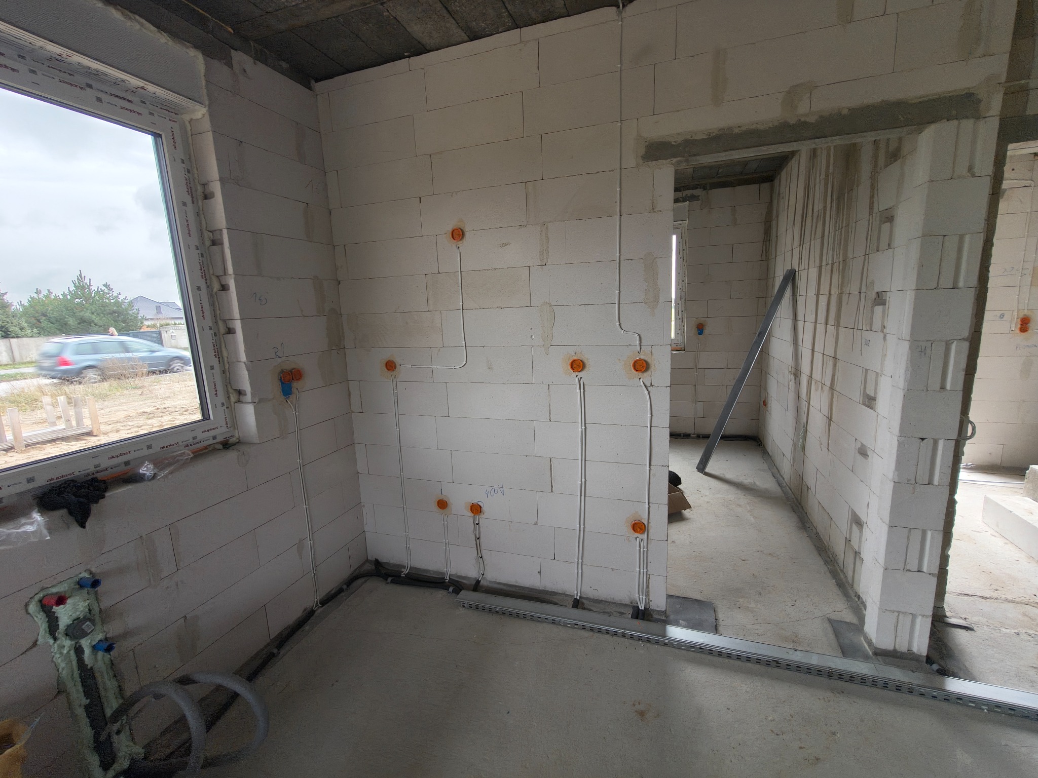 Wnętrze w trakcie prac elektrycznych: widoczne kable w peszlach prowadzone po ścianach z betonu komórkowego, puszki instalacyjne. Surowy stan, okno z widokiem na zewnątrz.