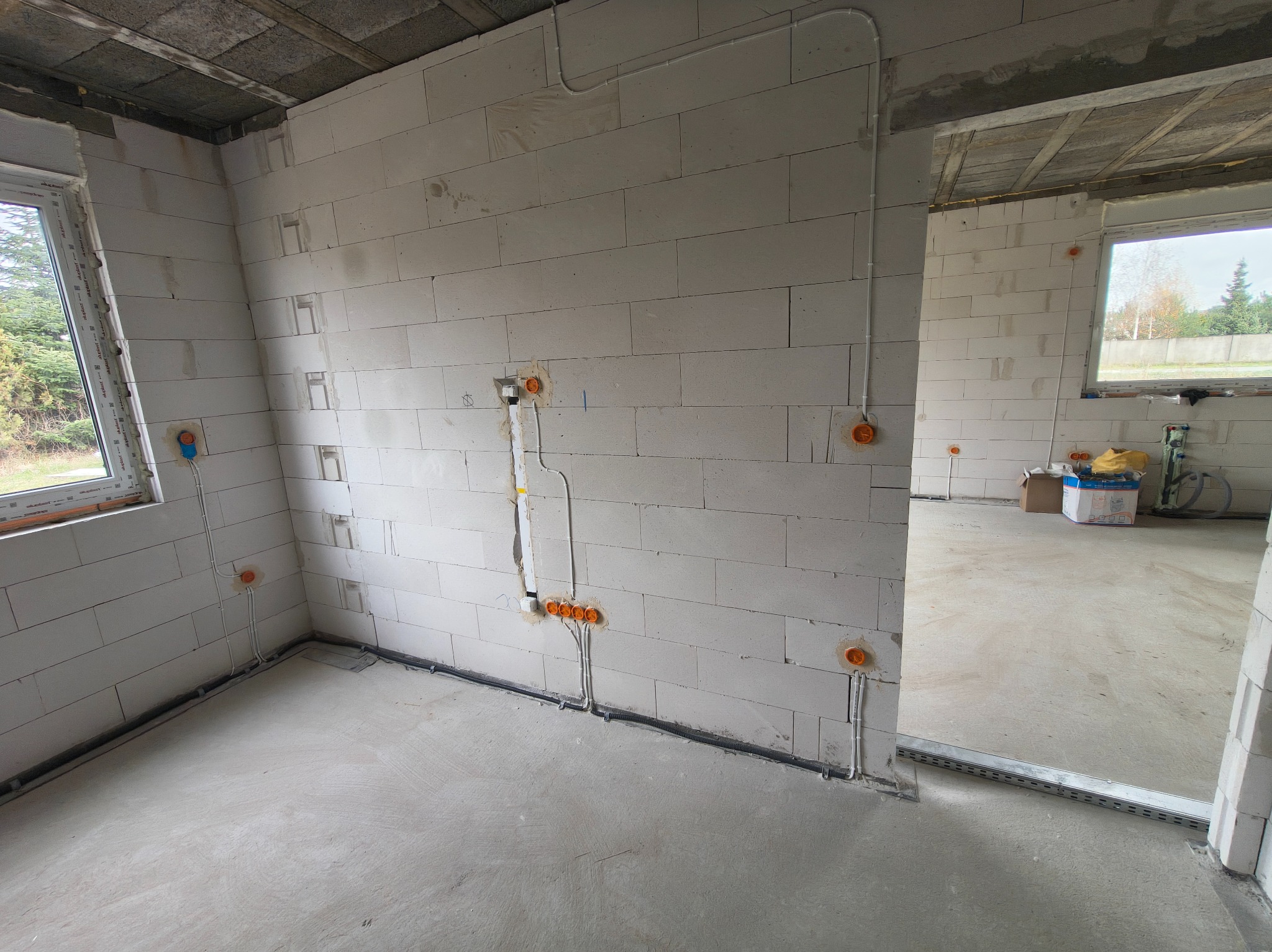 Surowy stan: wnętrze z instalacją elektryczną w ścianach z betonu komórkowego, widoczne puszki i peszle. Podłoga betonowa, okno z widokiem na zieleń.