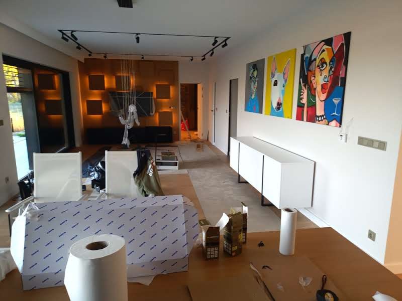 Nowoczesny salon w trakcie wykańczania: oświetlenie szynowe, biała komoda pod obrazami, elementy zabezpieczone folią. Widoczne prace wykończeniowe.