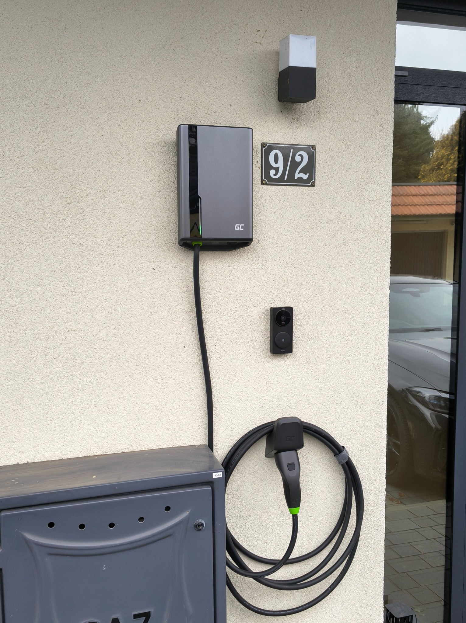 Szara stacja ładowania pojazdów elektrycznych GC zamontowana na elewacji domu obok skrzynki gazowej i numeru 9/2, z podłączonym kablem i wtyczką. Widoczny fragment samochodu.