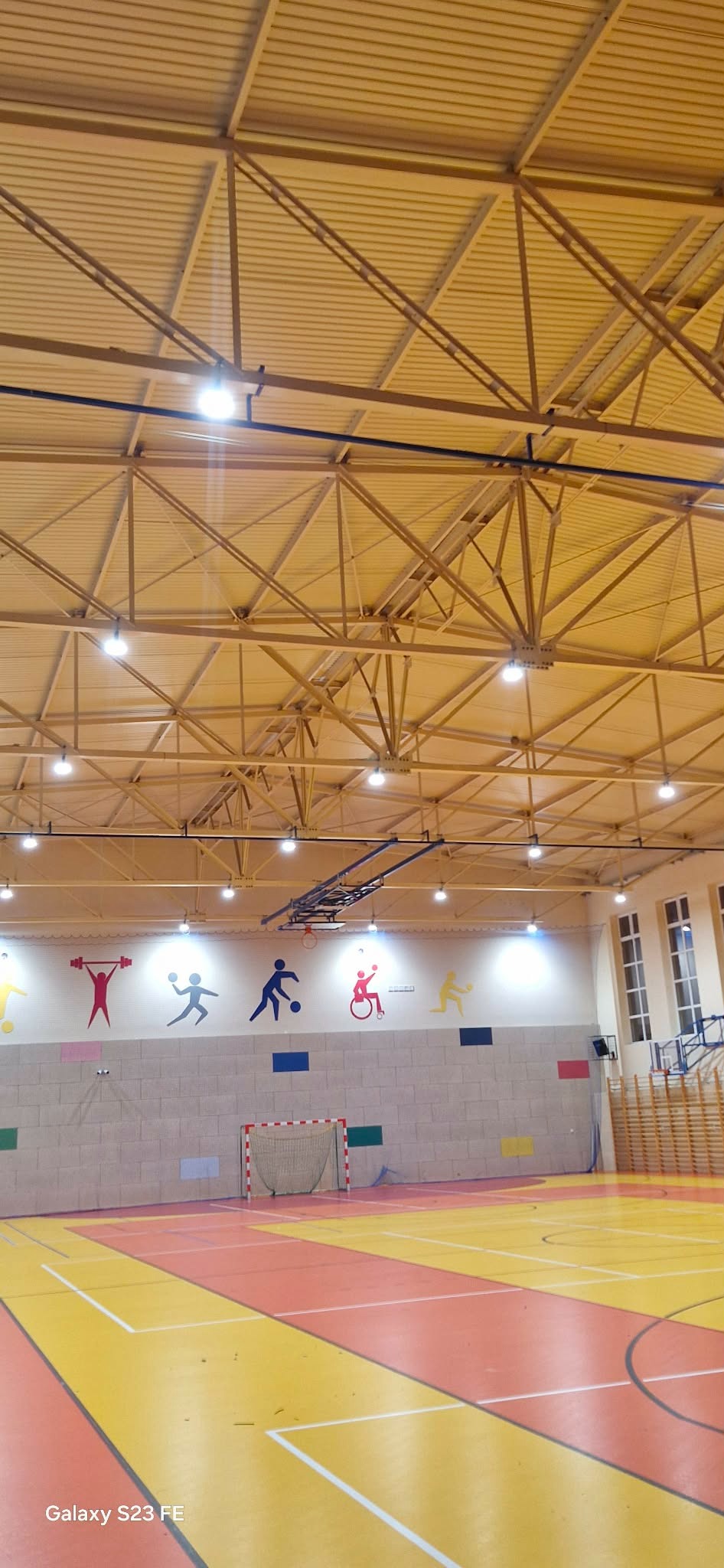 Oświetlenie hali sportowej z widoczną konstrukcją dachu, lampami i ścianą z kolorowymi sylwetkami sportowców. Na podłodze bramka do piłki ręcznej.