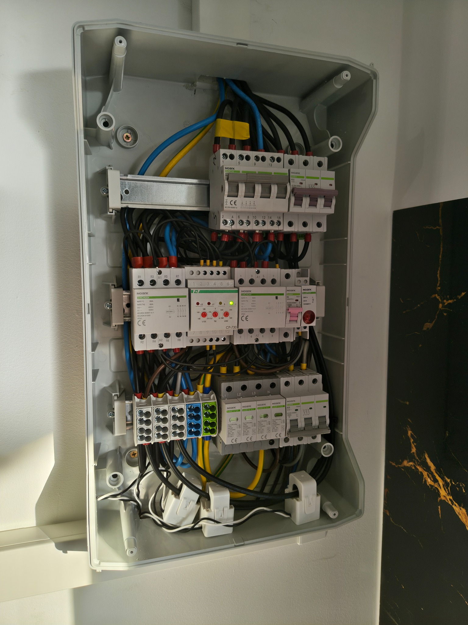 Otwarta skrzynka elektryczna z bezpiecznikami, przewodami w kolorach niebieskim, żółtym i czarnym. Widoczne elementy montażowe i zabezpieczenia.