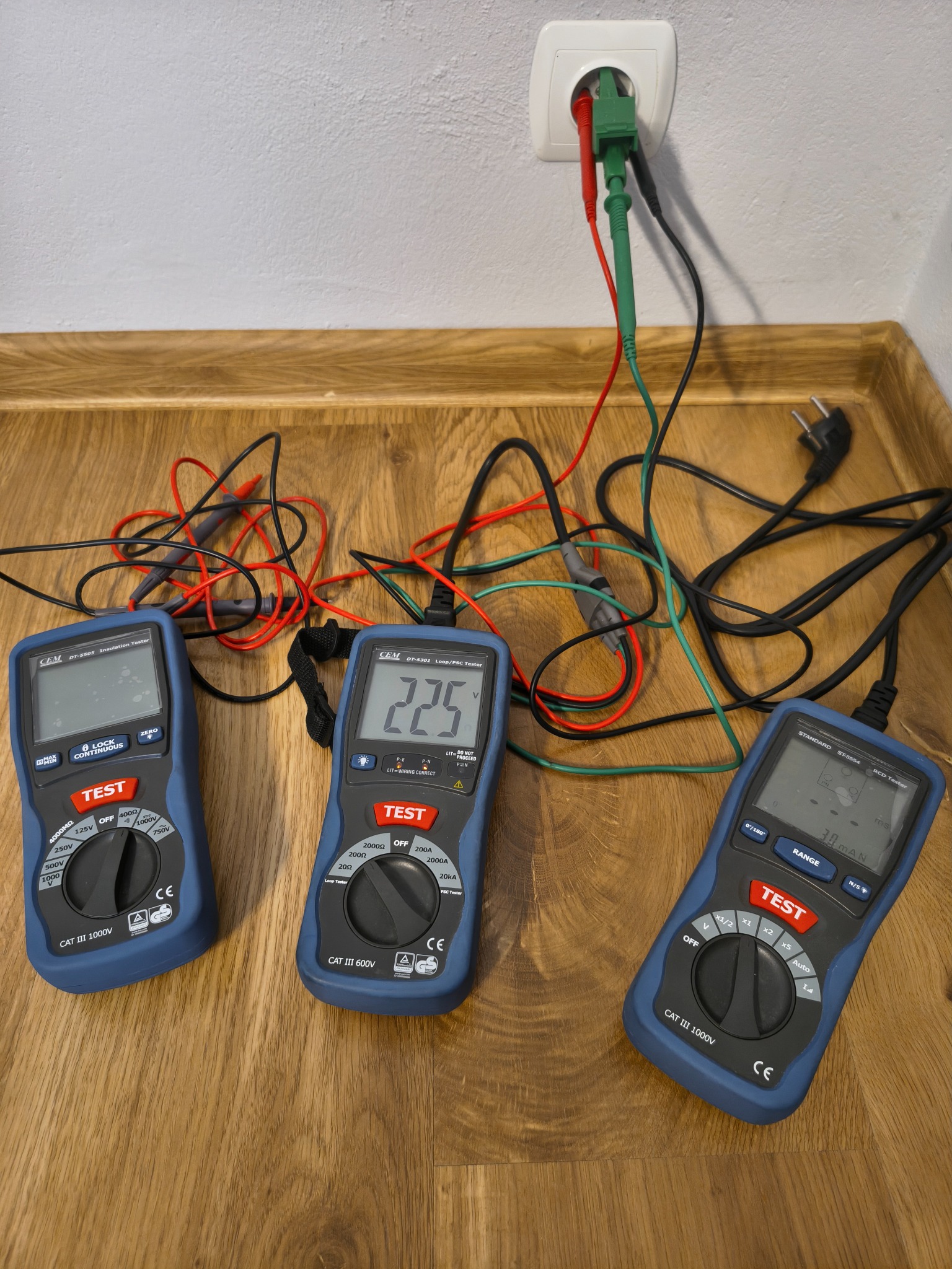 Trzy multimetry CEM z podłączonymi przewodami pomiarowymi, jeden wpięty do gniazdka. Mierniki leżą na drewnianej podłodze. Widoczne wartości pomiarowe na wyświetlaczach.
