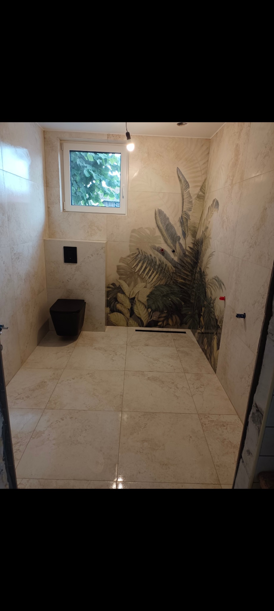 Wykończona łazienka z beżowymi płytkami, czarną toaletą podwieszaną i motywem tropikalnych liści na ścianie. Widoczne okno. Całość utrzymana w jasnej tonacji.