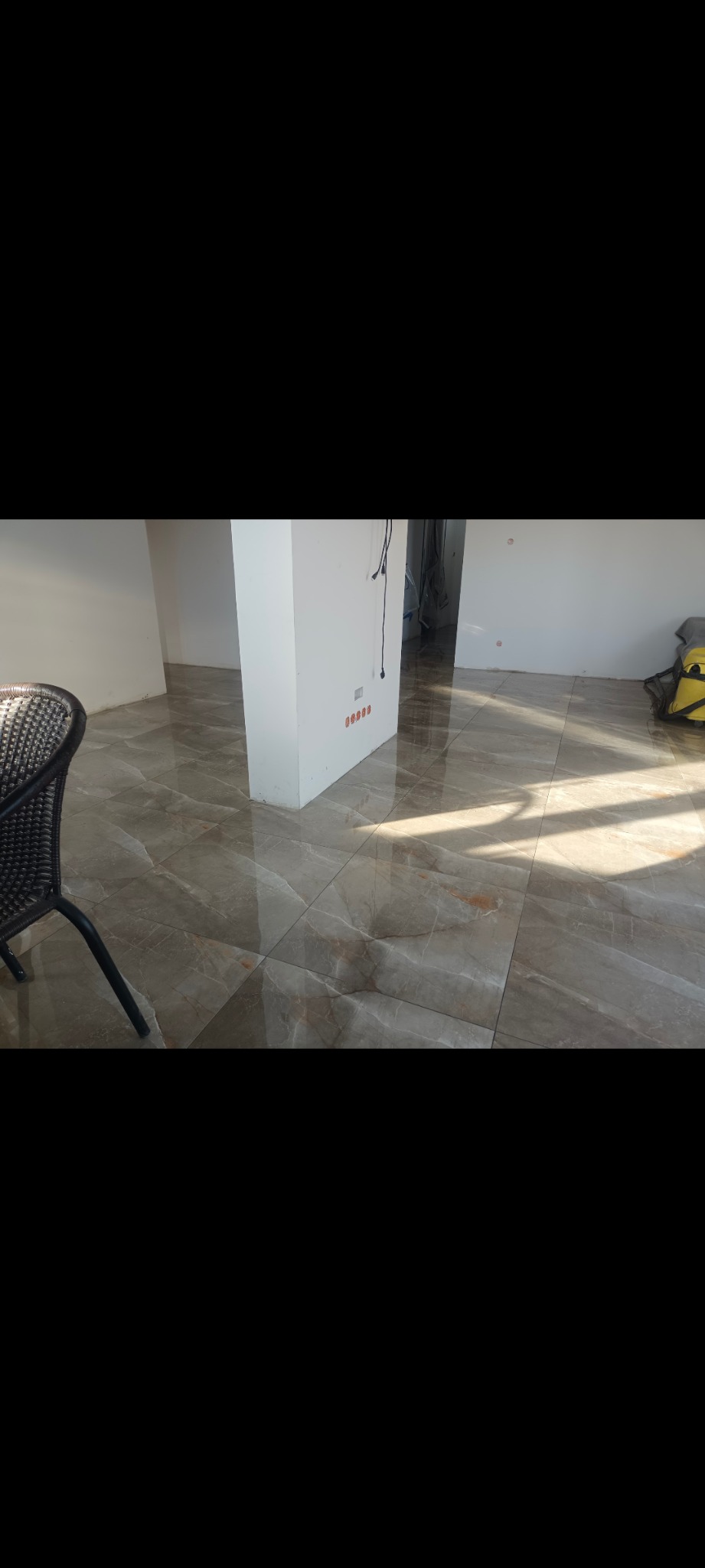 Wykończona podłoga z płytek imitujących marmur w jasnym, przestronnym wnętrzu. Widoczne gniazdka elektryczne i fragment krzesła z rattanu.