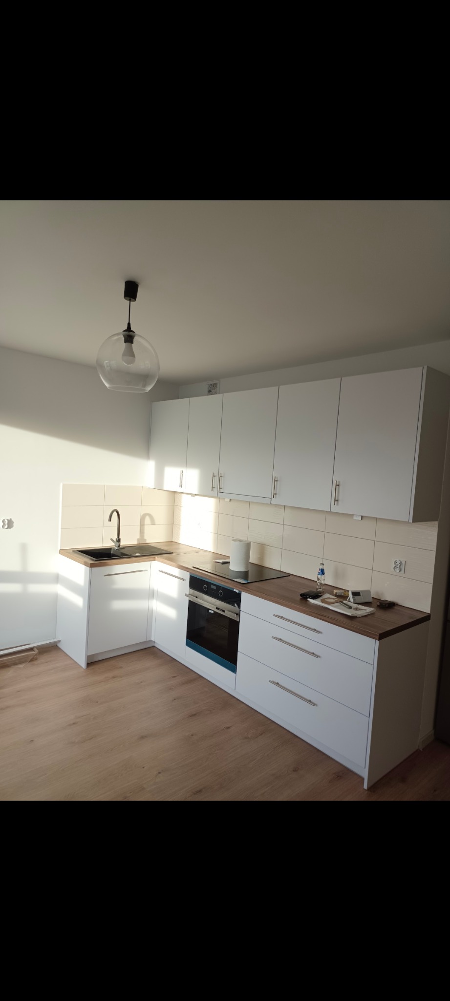 Jasna kuchnia z białymi szafkami, drewnianym blatem i panelami. Nad blatem białe płytki. Na blacie kuchenka indukcyjna i akcesoria. Nowoczesna lampa.