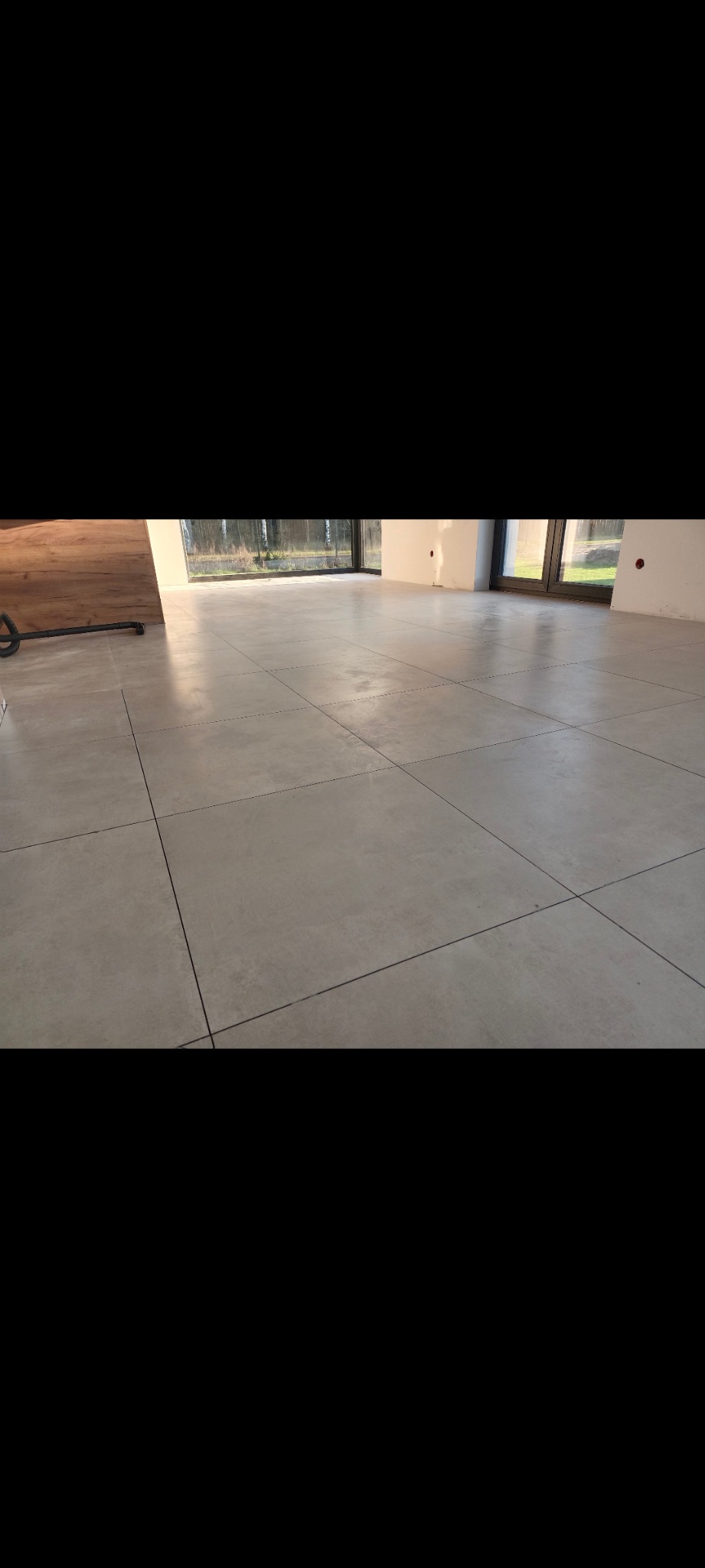 Jasnoszara podłoga z dużych kwadratowych płytek, widok na okno i fragment ściany z instalacją elektryczną. Minimalistyczne wykończenie wnętrza.