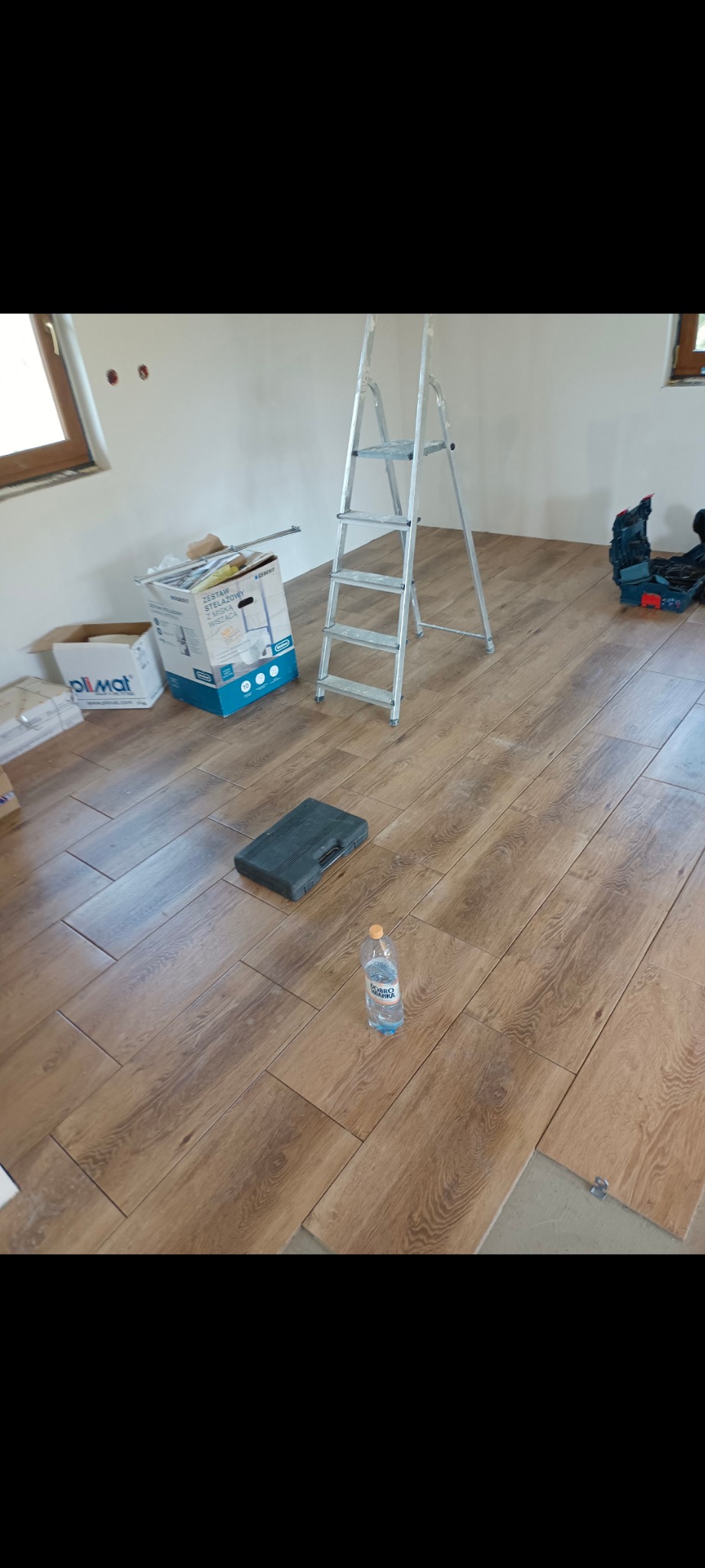 Układanie podłogi z płytek imitujących drewno. Widoczna drabina, narzędzia, kartony z materiałami i butelka wody. Częściowo ułożona podłoga w jasnym pomieszczeniu.