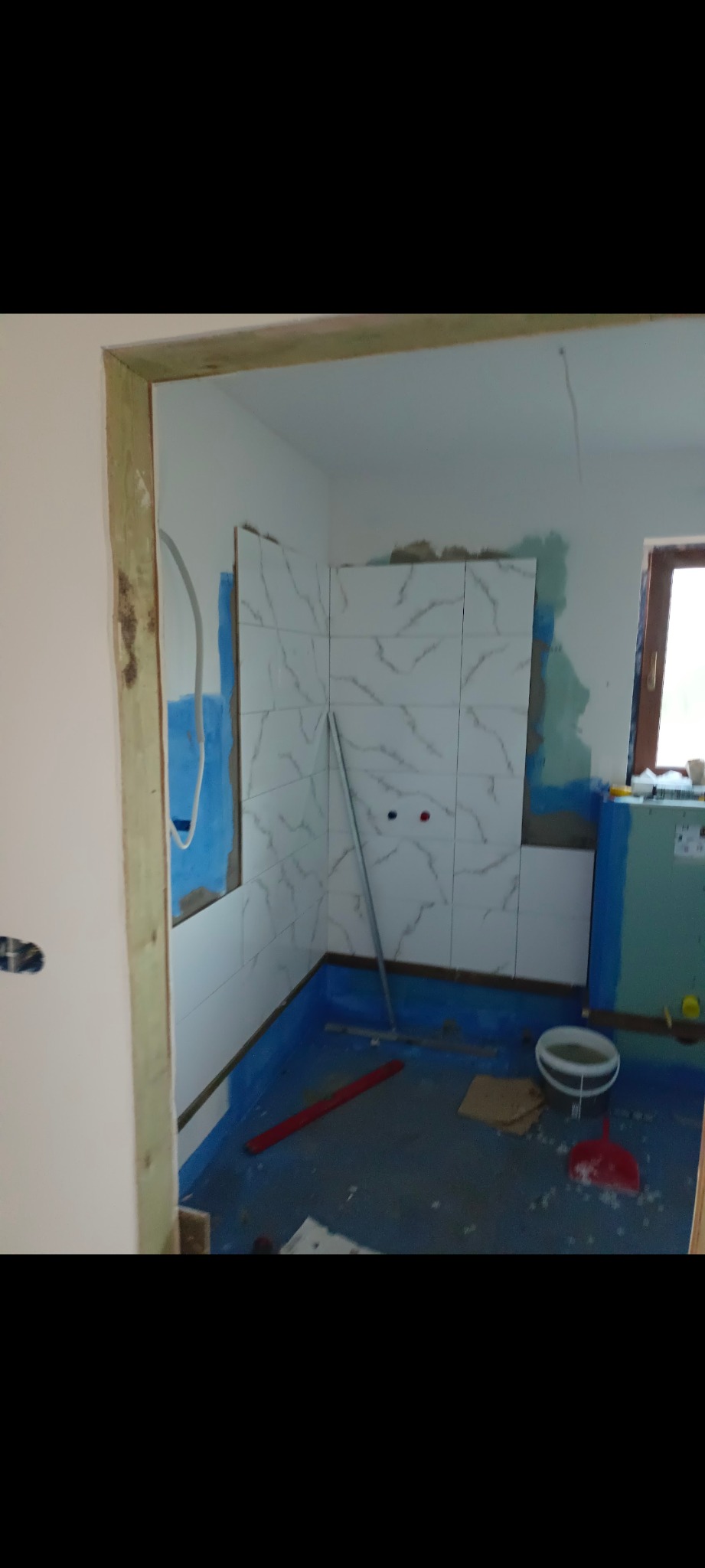 Łazienka w trakcie remontu: białe płytki marmurowe na ścianach, niebieska hydroizolacja podłogi, widoczny otwór drzwiowy z surową ościeżnicą.