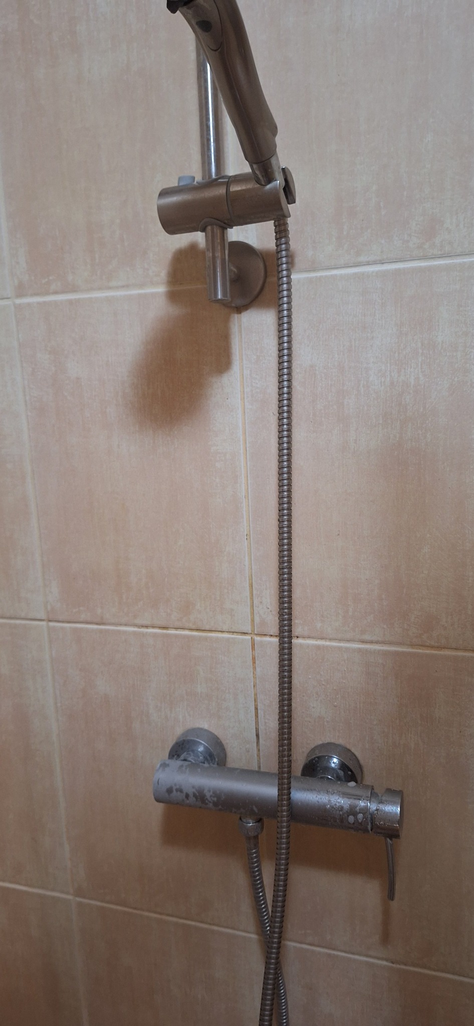 Z bliska: bateria prysznicowa z osadem kamiennym na beżowych płytkach. Widoczna słuchawka, wąż i uchwyt. Potrzeba czyszczenia lub wymiany.