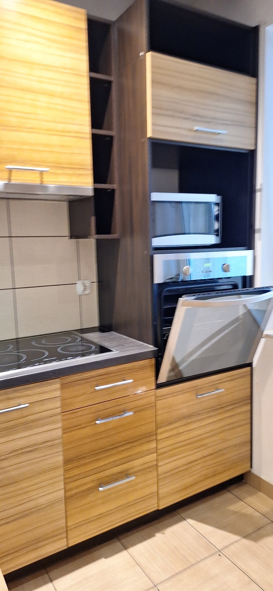 Częściowy widok czystej, nowoczesnej kuchni z drewnianymi szafkami, płytą grzewczą i otwartym piekarnikiem. Uwagę zwraca minimalistyczny design i dbałość o detale.