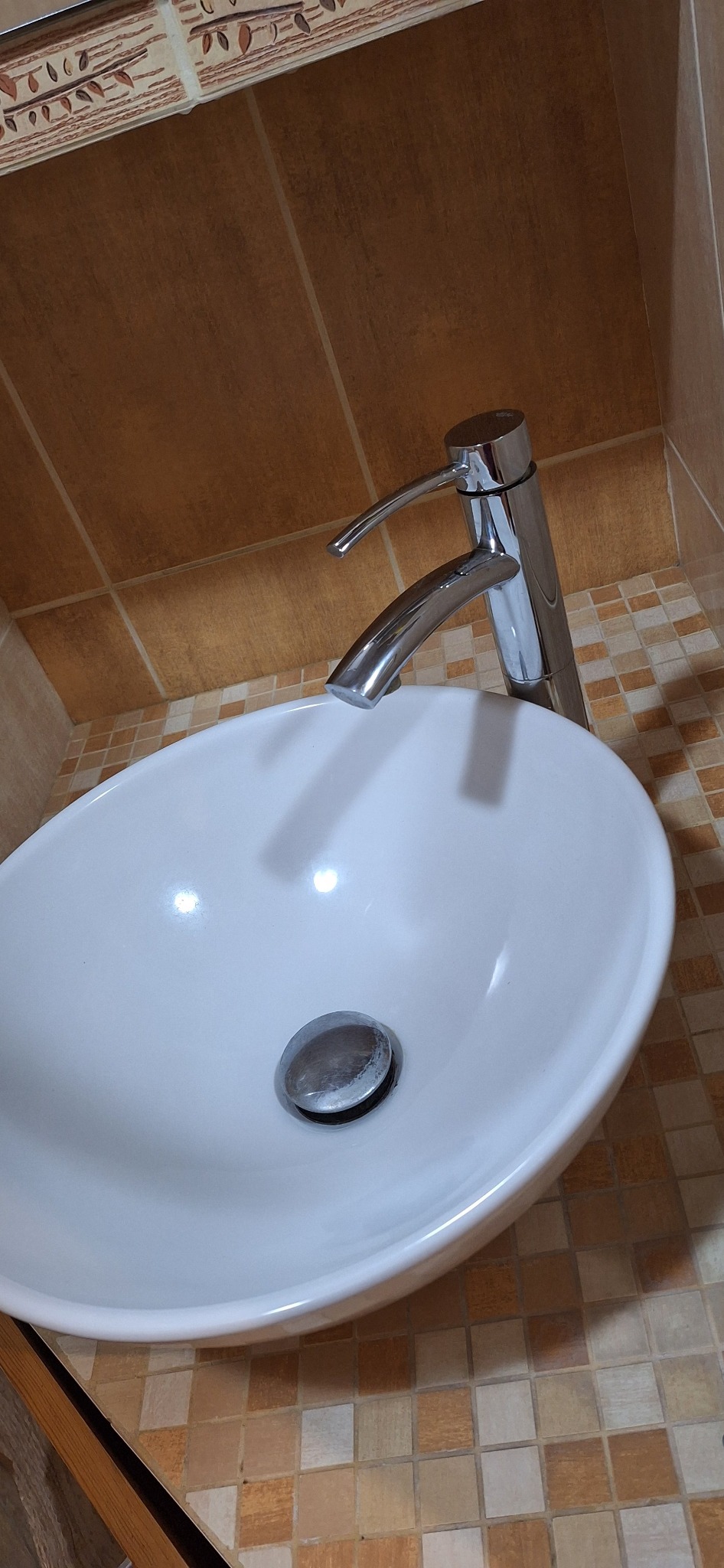 Biała umywalka z chromowaną baterią na tle mozaiki w łazience. Widoczne detale baterii i odpływu. Ujęcie z góry, podkreślające czystość i estetykę.