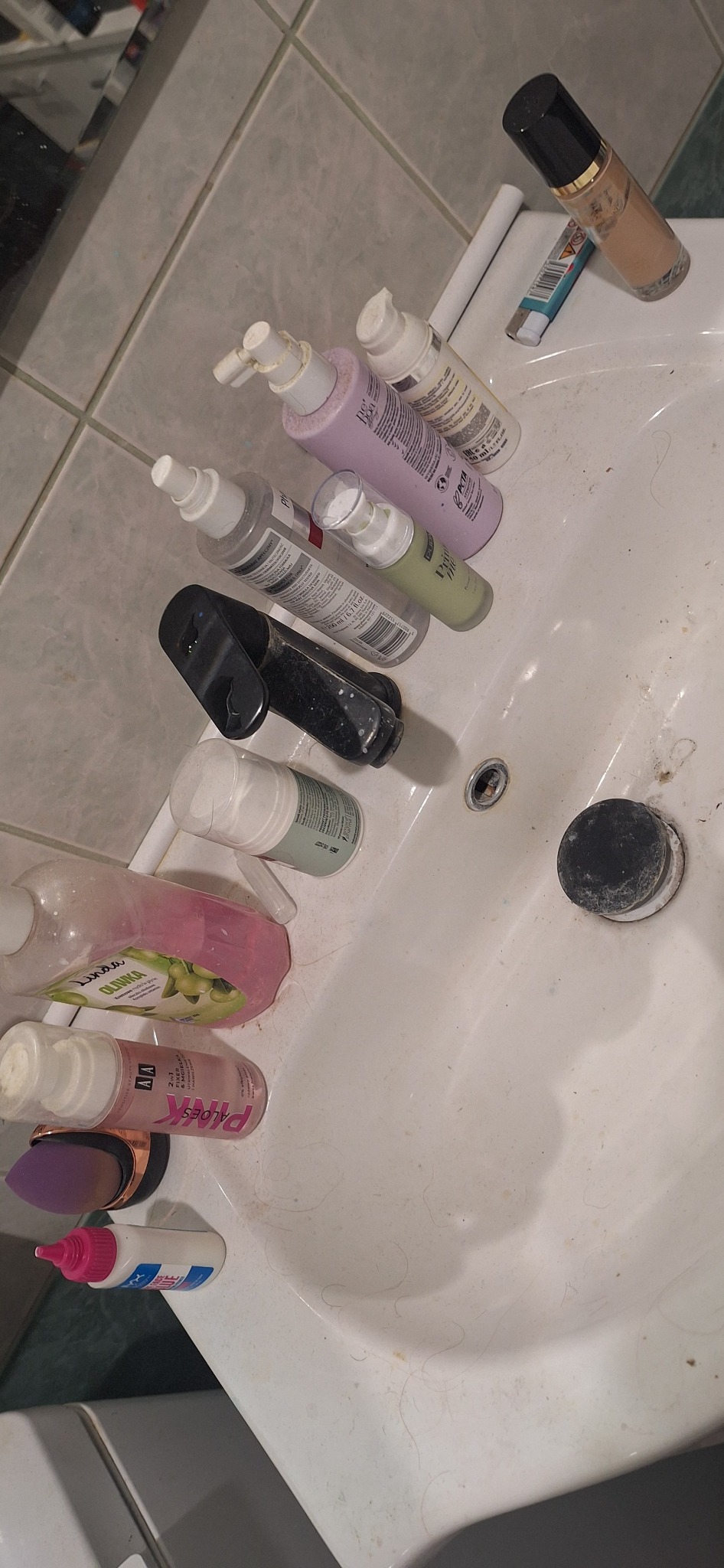 Zanieczyszczona umywalka z kosmetykami i włosami. Widoczne ślady użytkowania i brak czystości. Potrzeba gruntownego sprzątania łazienki.