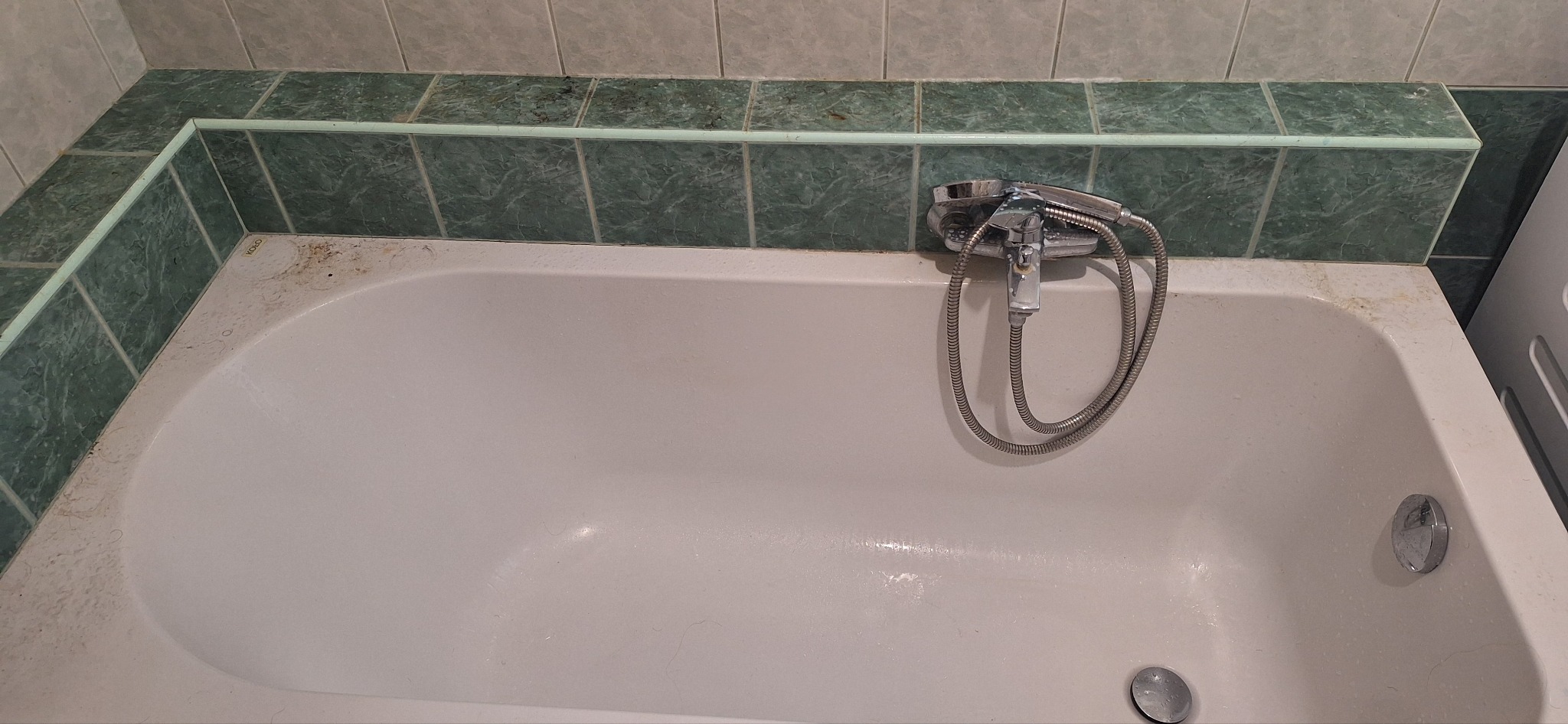 Zaniedbana wanna z osadami i brudnymi fugami, widoczna słuchawka prysznicowa. Potrzeba gruntownego czyszczenia, aby przywrócić jej blask.