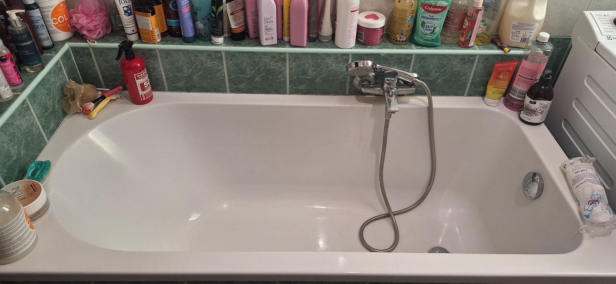 Biała wanna z chromowaną baterią i prysznicem, otoczona kosmetykami na zielonej półce. Widoczne ślady użytkowania, sugerujące potrzebę sprzątania.