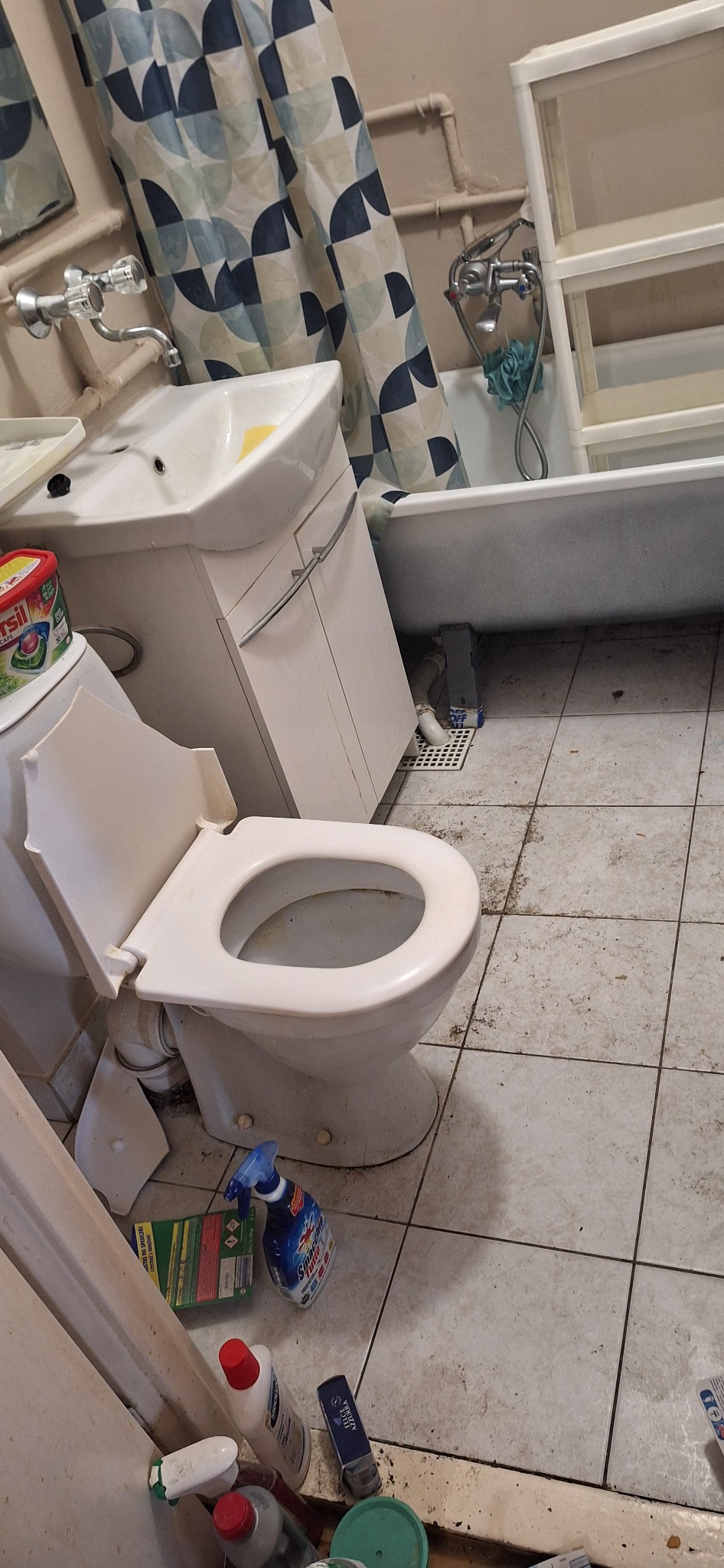 Zaniedbana łazienka z widocznymi zabrudzeniami na podłodze i toalecie. Środki czystości leżące na podłodze sugerują potrzebę gruntownego sprzątania.