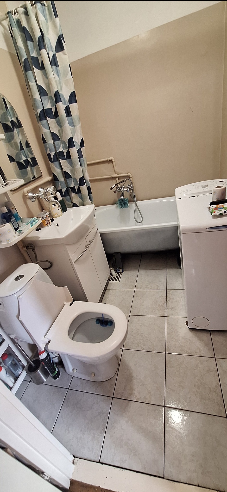 Umywalka, wanna, toaleta i pralka w łazience z beżowymi ścianami i szaro-beżowymi płytkami. Nad wanną niebiesko-biała zasłonka. Widok z góry.