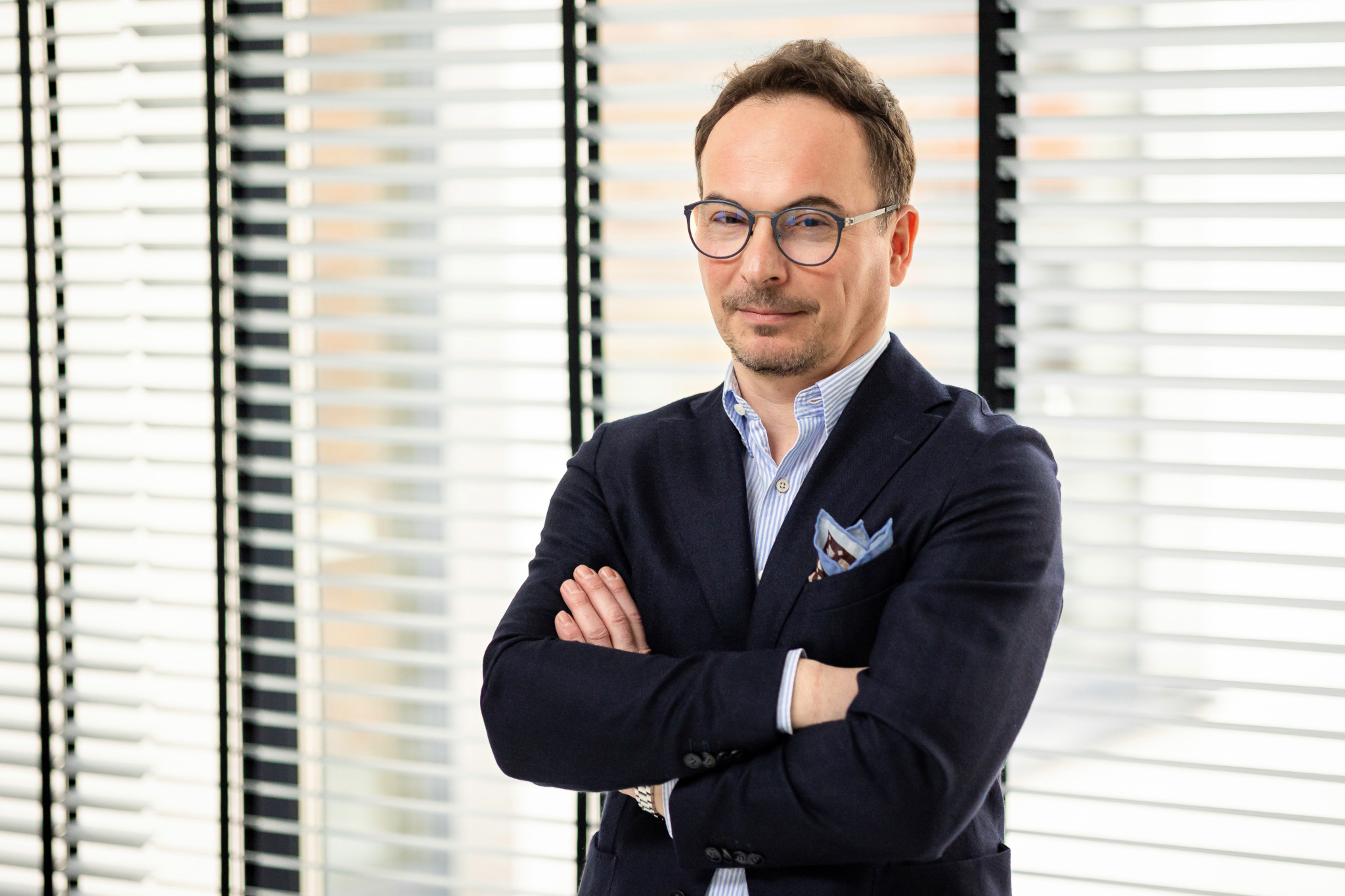 Portret mężczyzny w okularach, ubrany w marynarkę, z rękami skrzyżowanymi na tle żaluzji. Profesjonalny wizerunek trenera lub rekrutera.