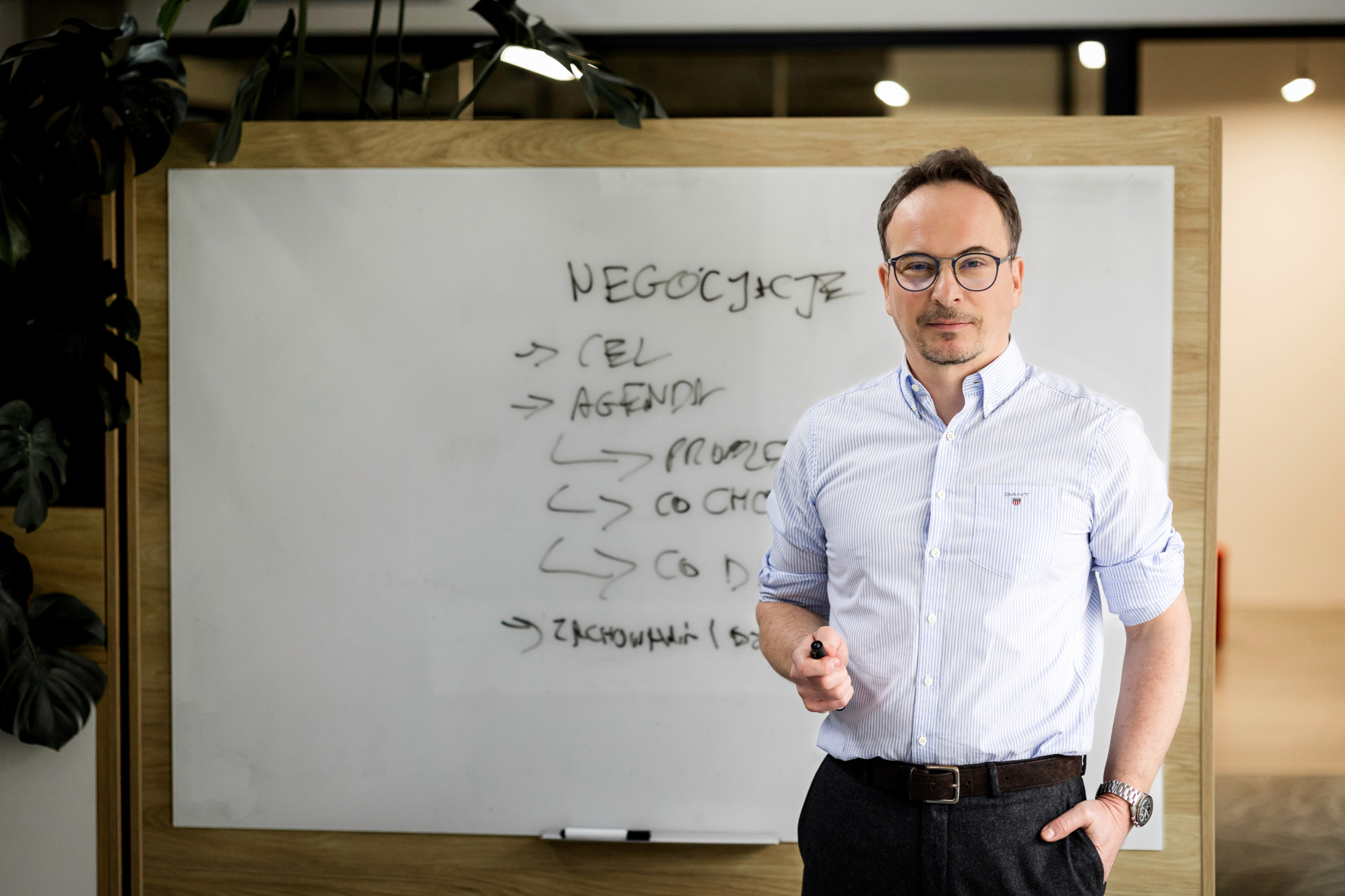 Mężczyzna w okularach prezentuje na białej tablicy zagadnienia negocjacji, trzymając marker. Widoczne etapy: cel, agenda, profile, co chcę, co dam.