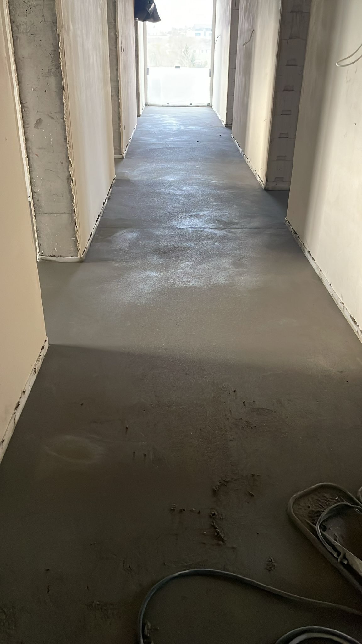 Świeżo wylana, szara posadzka jastrychowa w długim korytarzu, widok z dołu, z widocznymi śladami po narzędziach i fragmentem wiaderka w rogu kadru.