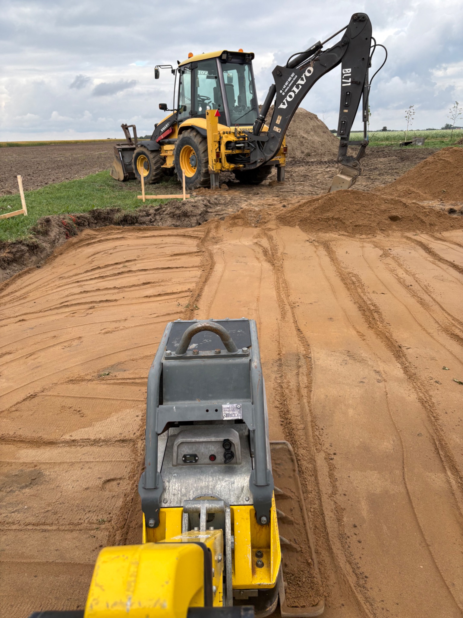 Żółta zagęszczarka wibrująca ubija piasek, w tle koparka Volvo i drewniane znaczniki wykopu na polu. Prace ziemne w toku.