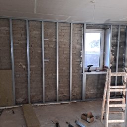 Man-bud - Montaż metalowej konstrukcji na ceglanej ścianie, częściowo ocieplonej wełną mineralną. Widoczne okno, drabina i narzędzia. Przygotowanie do wykończenia ścian.