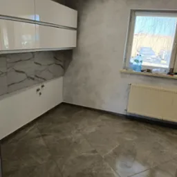 Wykończona łazienka z szarymi płytkami podłogowymi imitującymi marmur, białe szafki wiszące i ściany z efektem dekoracyjnym. Grzejnik pod oknem.