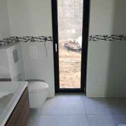 Man-bud - Nowoczesna łazienka z umywalką, toaletą i mozaiką. Widoczne przyłącza hydrauliczne i okno z widokiem na plac budowy. Minimalistyczny design, szare płytki podłogowe.