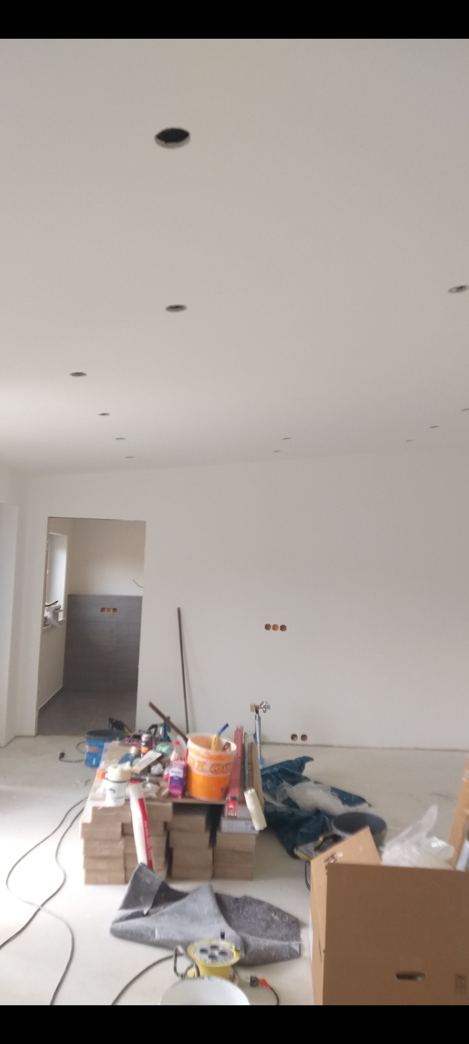 Wnętrze w trakcie prac wykończeniowych: białe ściany, sufit z otworami na oświetlenie, narzędzia i materiały budowlane zgromadzone w pomieszczeniu.