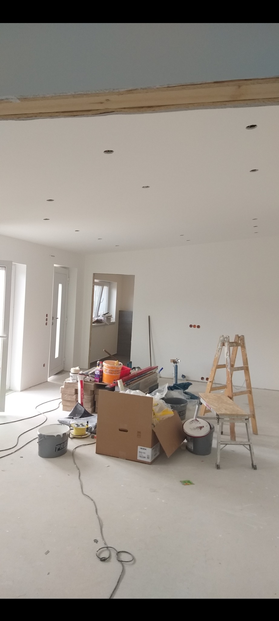 Jasne wnętrze w trakcie prac wykończeniowych: białe ściany, sufit z punktowym oświetleniem, drabina i akcesoria malarskie na podłodze. Widok na sąsiednie pomieszczenie.