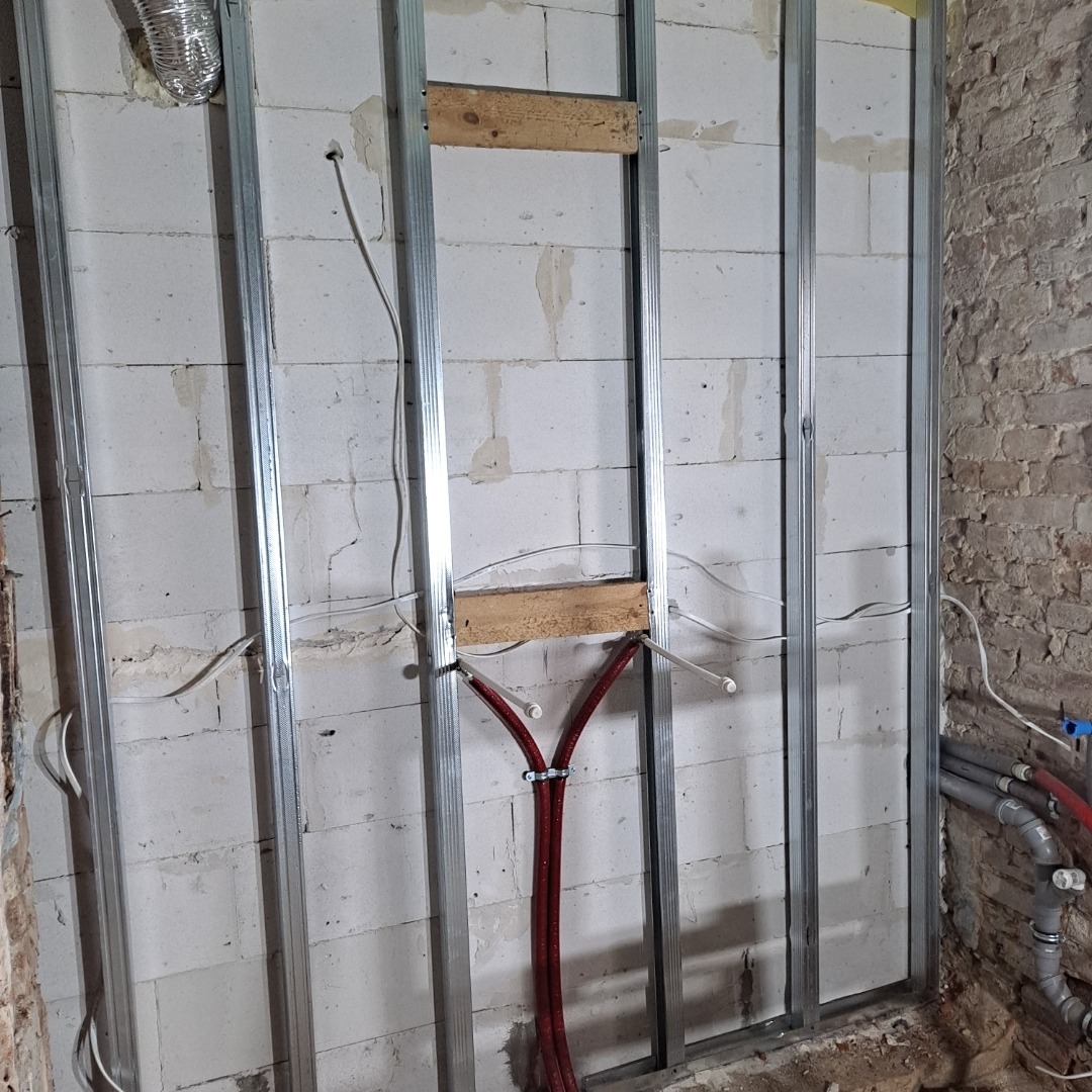 Ściana z bloków, z metalową konstrukcją, drewnianymi elementami i instalacją wodną w trakcie montażu. Widoczne przewody i rury. Surowy stan wykończenia.