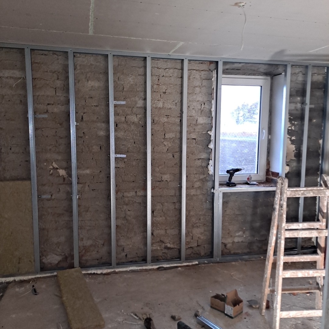 Montaż metalowej konstrukcji na ceglanej ścianie, częściowo ocieplonej wełną mineralną. Widoczne okno, drabina i narzędzia. Przygotowanie do wykończenia ścian.