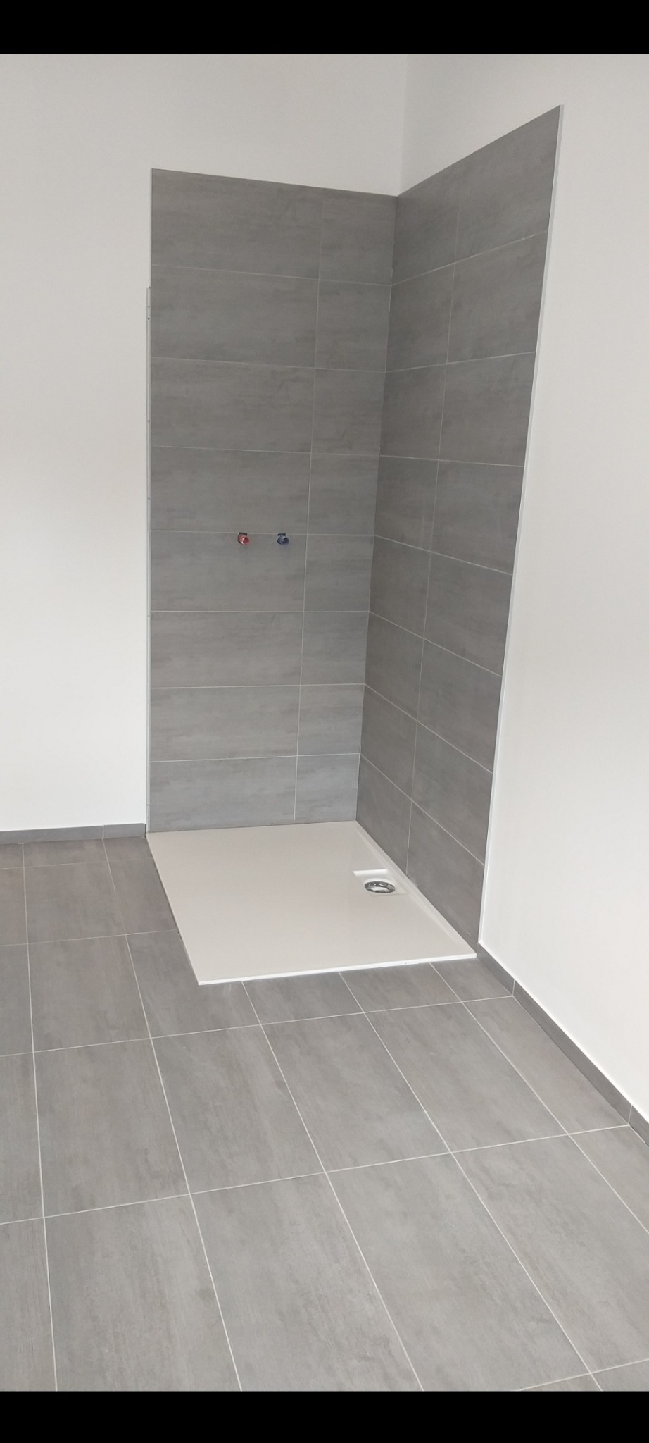 Wykończona łazienka w Żmigrodzie: szare płytki podłogowe i ścienne, biały brodzik. Minimalistyczny design, widoczne przyłącza wodne. Nowoczesne wnętrze.