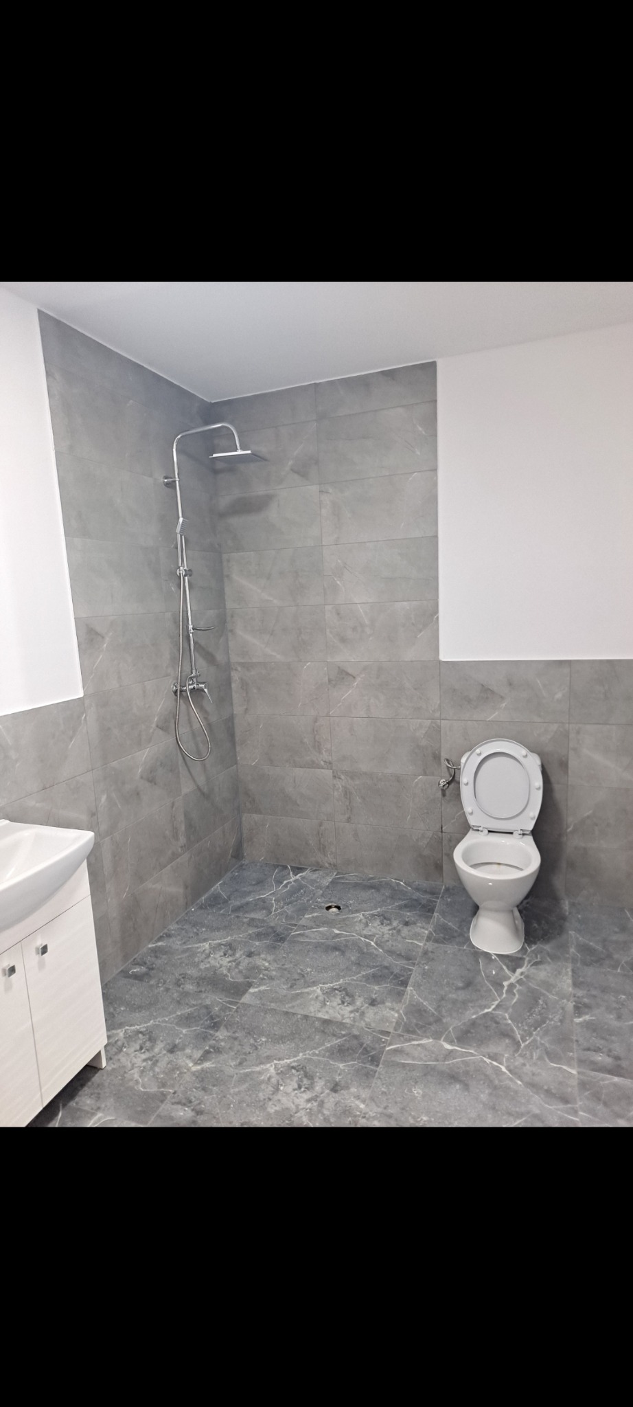 Wykończona łazienka w szarościach: marmurowe płytki, kabina prysznicowa typu walk-in, biała umywalka z szafką i toaleta. Minimalistyczny design, nowoczesne wykończenie.