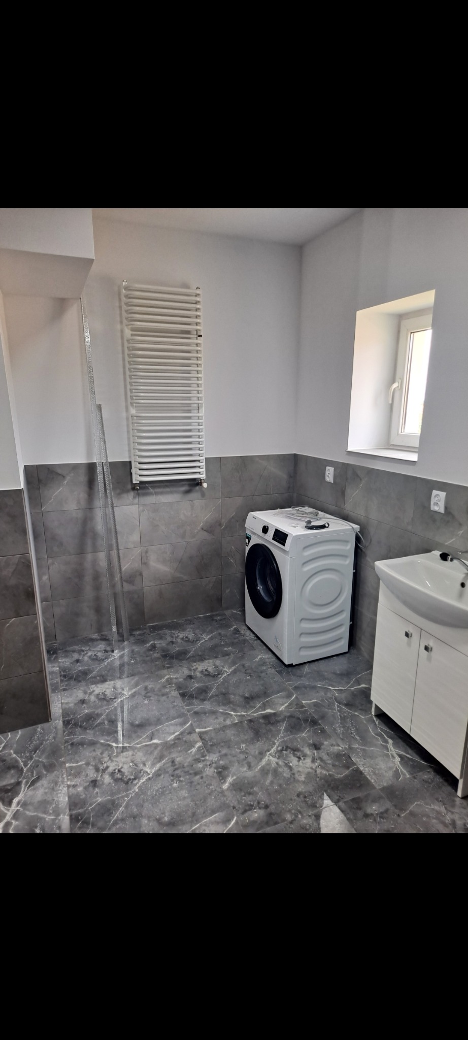 Wykończona łazienka z szarymi płytkami imitującymi marmur na podłodze i ścianach, białą pralką oraz grzejnikiem drabinkowym. Umywalka z szafką i okno.