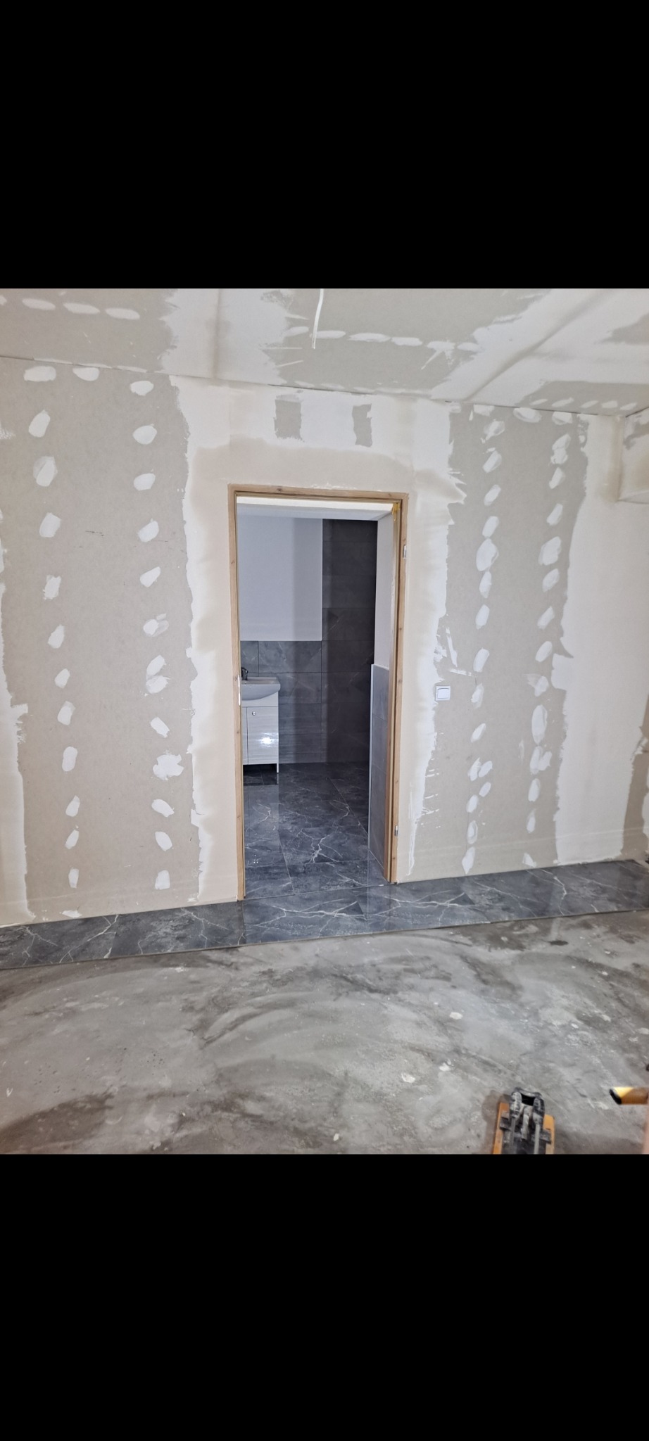Pokój w trakcie prac wykończeniowych: ściany z płyt gipsowo-kartonowych, widoczne łączenia, w tle wykończona łazienka z szarą glazurą i białą szafką umywalkową.