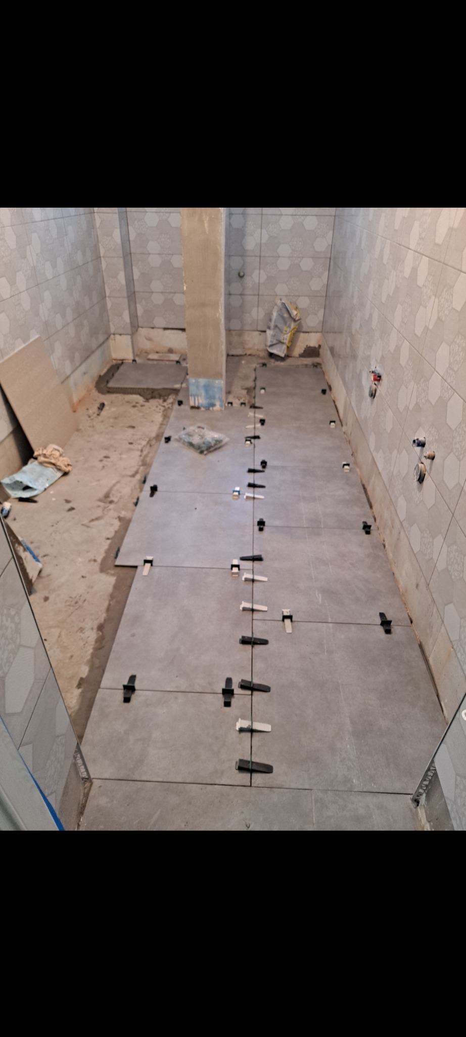 Układanie szarych płytek podłogowych w łazience, z użyciem klipsów poziomujących. Widoczne płytki ścienne z wzorem, przygotowane podłoże. Etap prac.