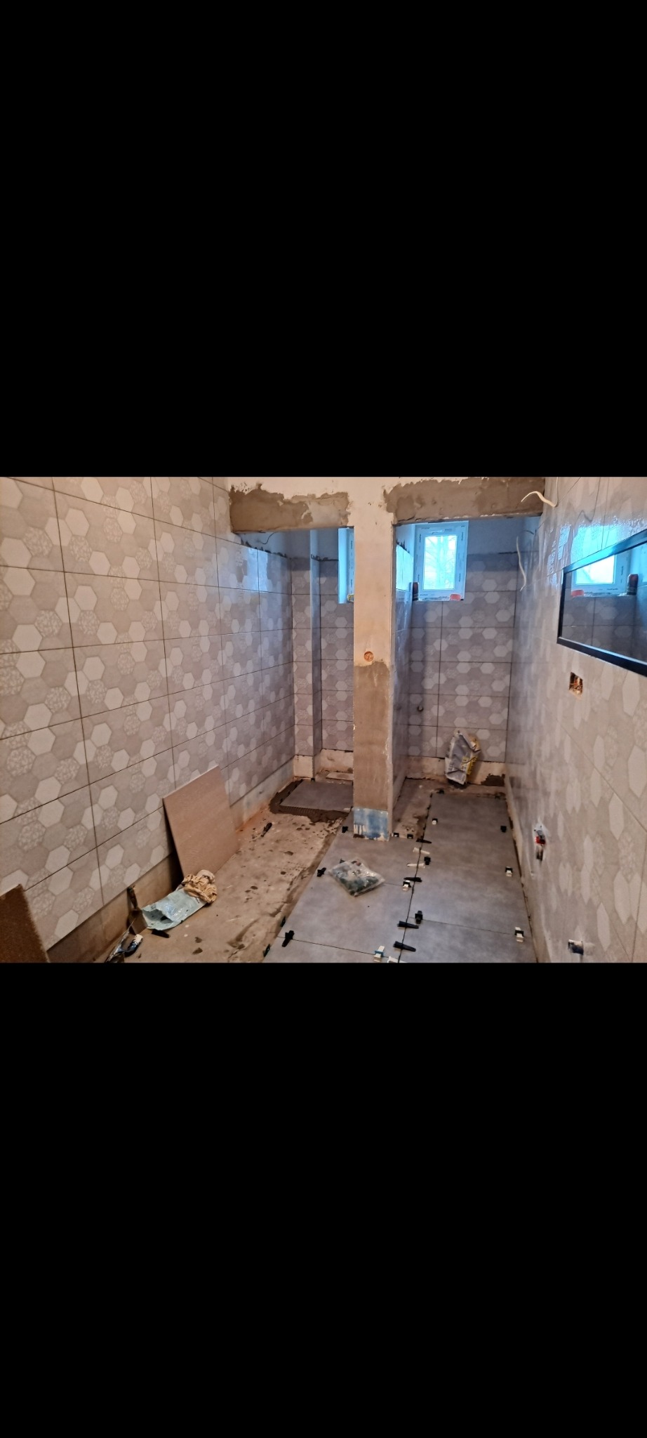 Wnętrze łazienki w trakcie prac. Na ścianach heksagonalne płytki, na podłodze szare płytki z krzyżykami dystansowymi. Widoczne okno i elementy instalacji.