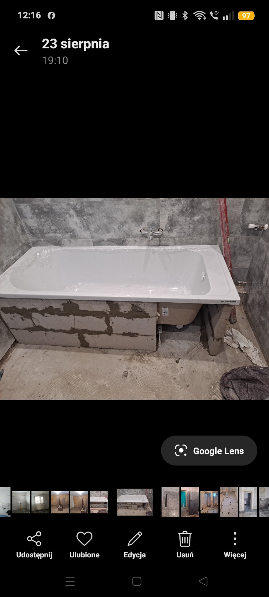 Biała wanna w trakcie obudowy z bloczków, szare płytki na ścianie, widoczny poziomica. Etap prac w łazience, przed wykończeniem.