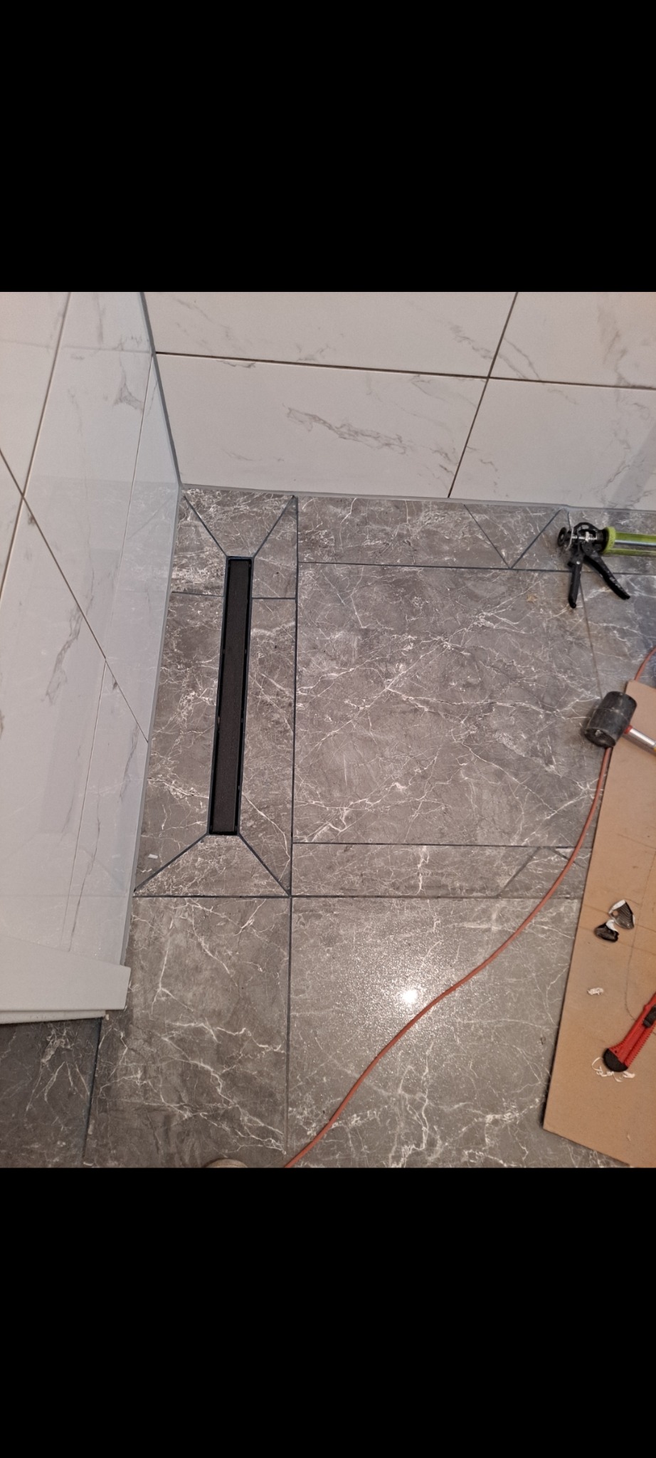 Ułożone płytki podłogowe w łazience z odpływem liniowym. Szare płytki imitujące marmur, białe płytki na ścianie. Widoczne narzędzia glazurnicze.