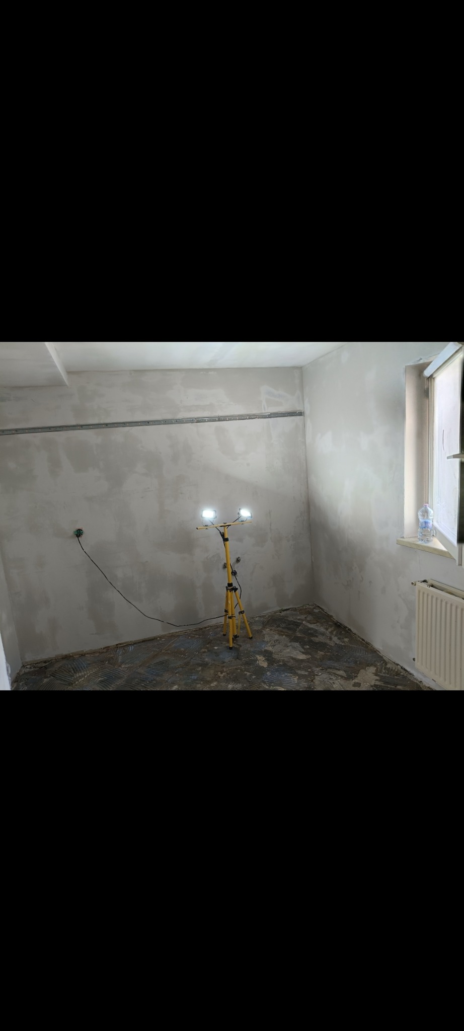 Pokój w trakcie prac przygotowawczych do wykończenia. Ściany po szpachlowaniu, podłoga częściowo zerwana. Żółty statyw z lampami oświetla wnętrze.