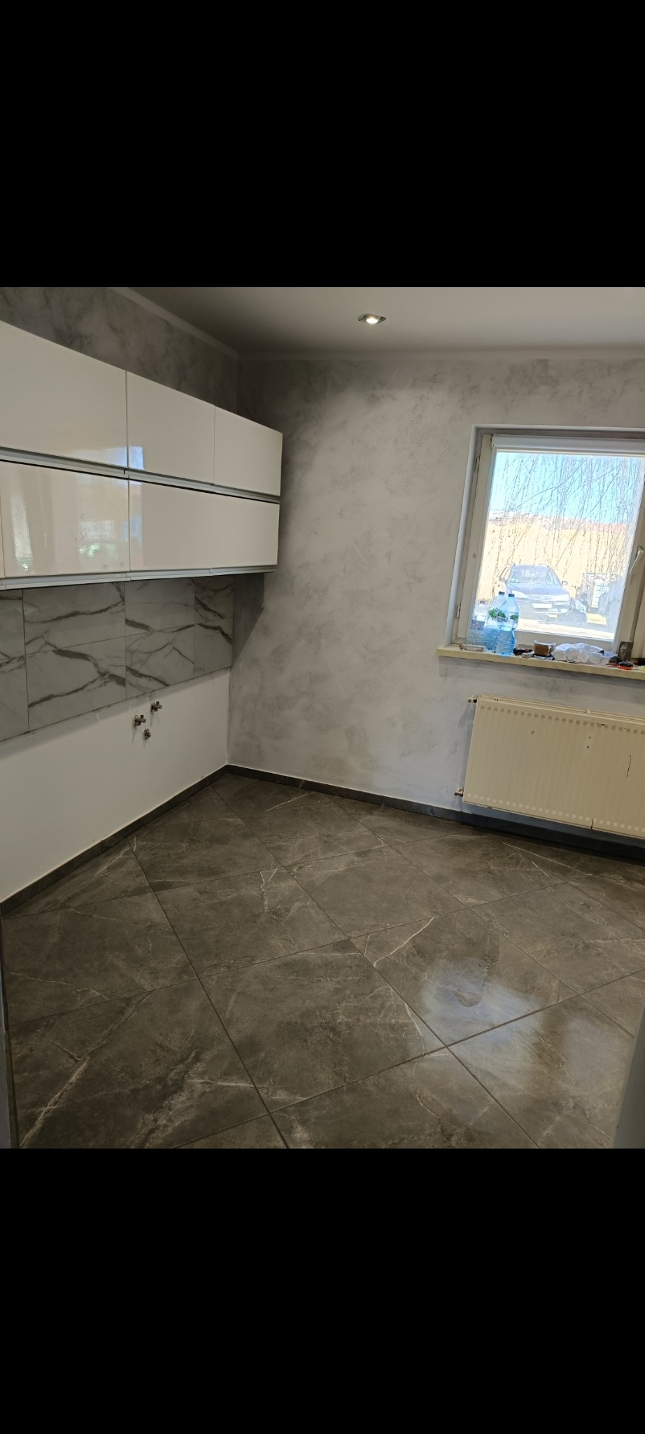 Wykończona łazienka z szarymi płytkami podłogowymi imitującymi marmur, białe szafki wiszące i ściany z efektem dekoracyjnym. Grzejnik pod oknem.