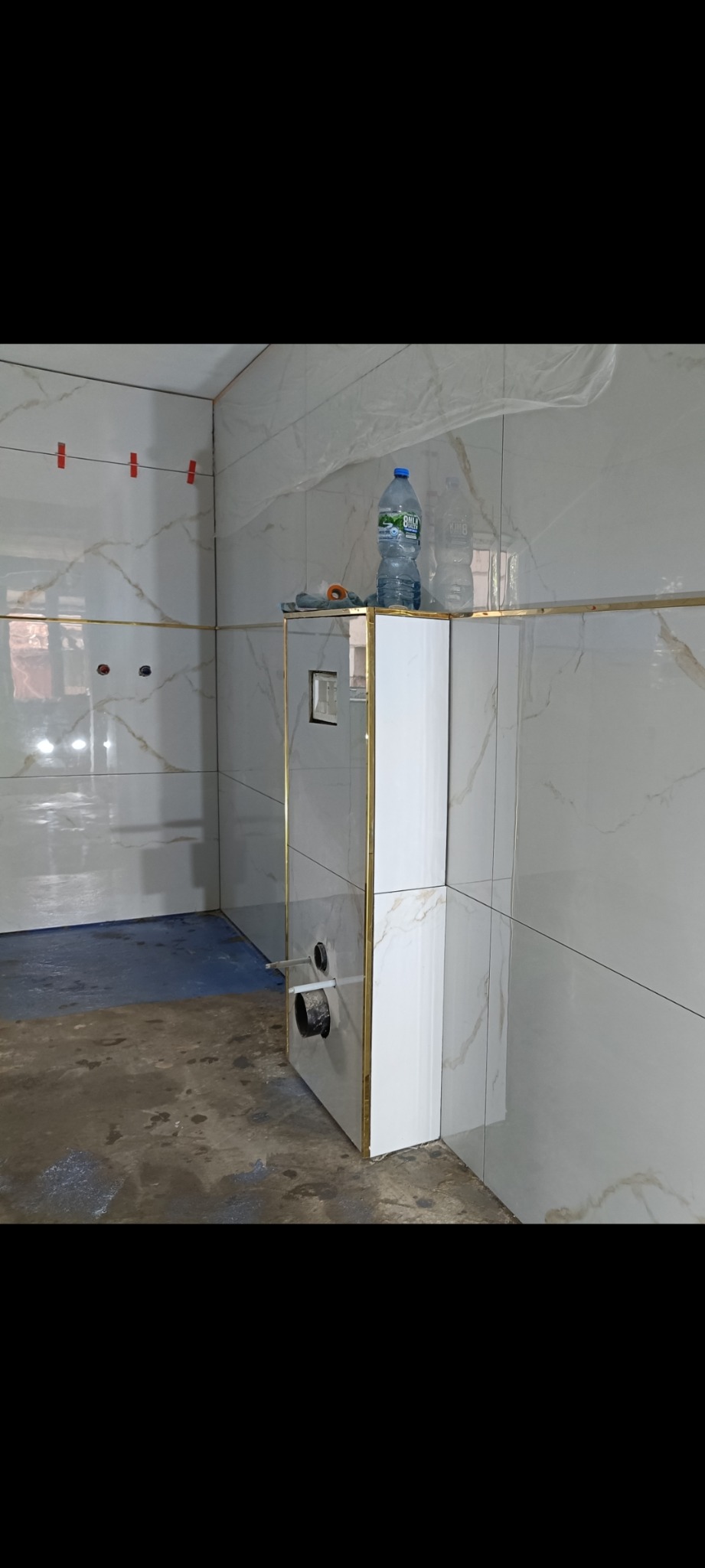 Wykończona obudowa WC z białych płytek z dekoracyjnymi złotymi listwami w łazience w trakcie remontu. Na obudowie stoi butelka wody.